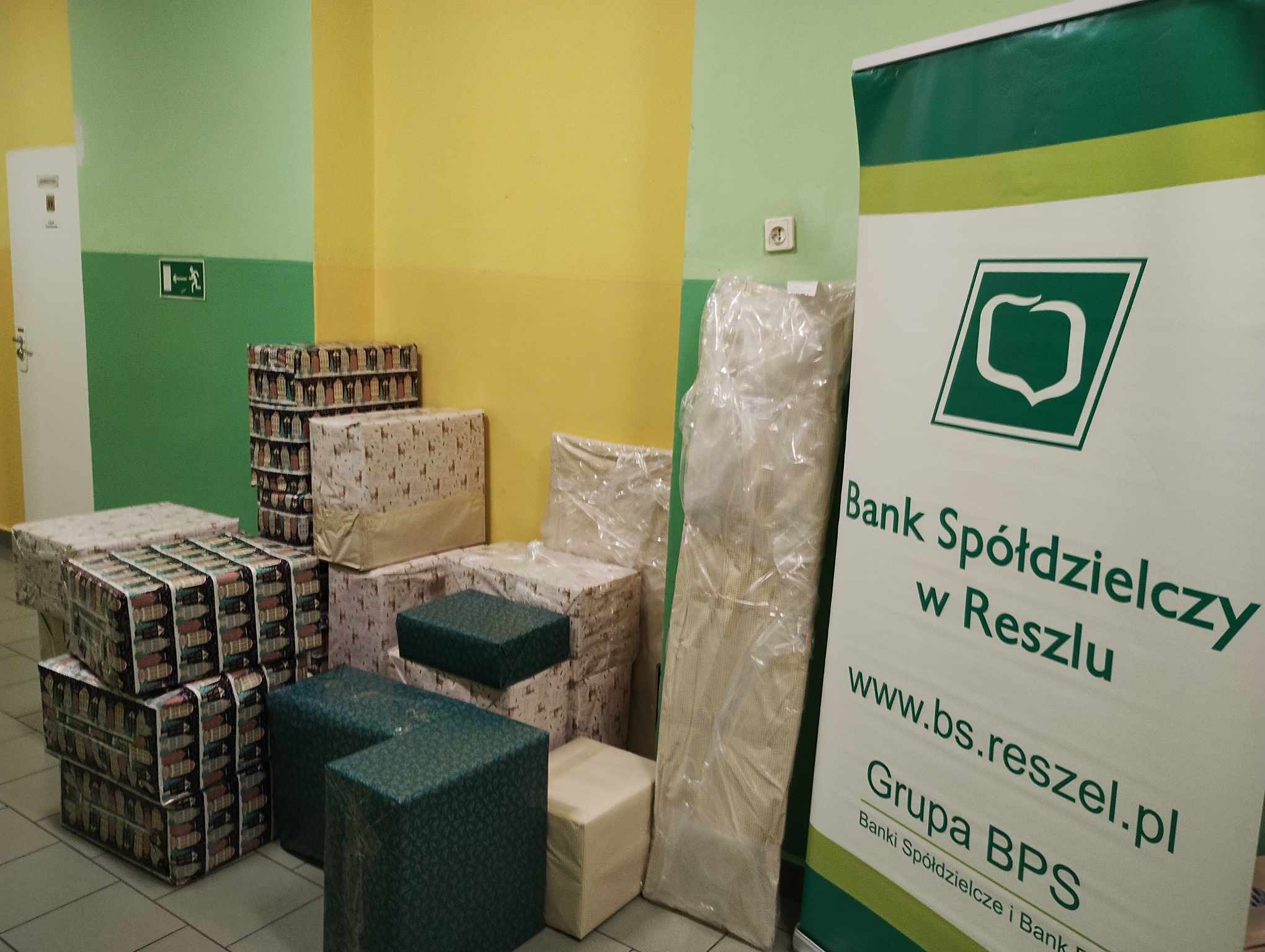 Stosy zapakowanych paczek obok baneru Banku Spółdzielczego w Reszlu